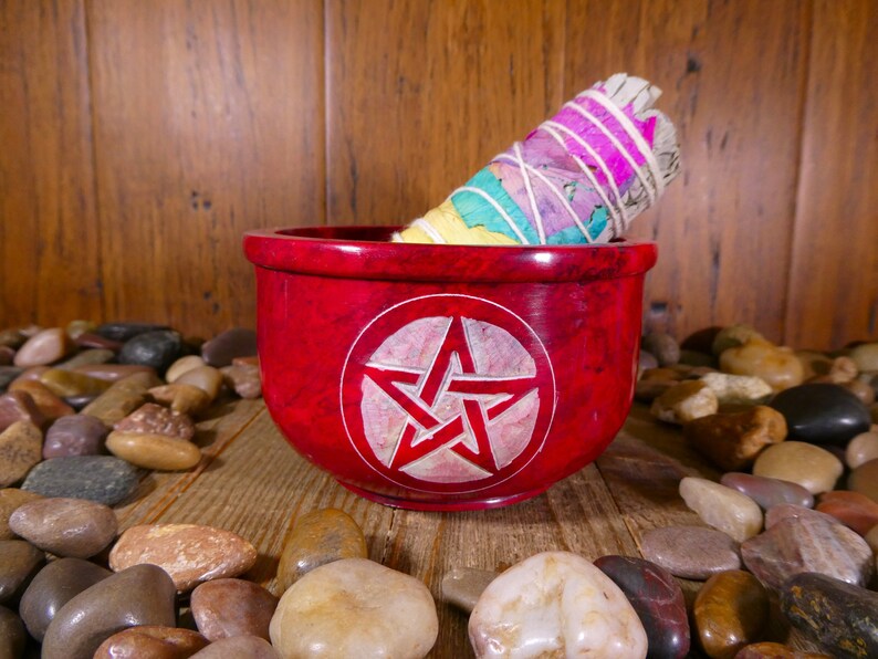 May include: A red, round, stone bowl with a white pentagram carved into the centre. A white sage smudge stick is resting on the rim of the bowl. The bowl is surrounded by smooth, grey and brown stones.