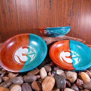 May include: Three round, wooden bowls with a teal and orange glaze. Each bowl features a white Om symbol carved into the surface.