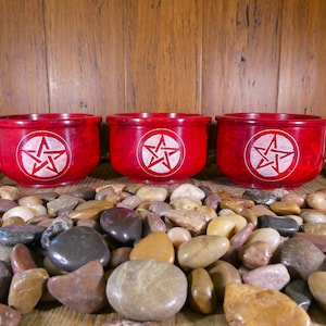 May include: Three red wooden bowls with a white pentagram design carved into the centre of each bowl. The bowls are sitting on a bed of small, smooth, multi-coloured stones.