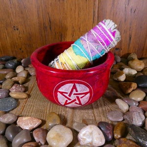 May include: A red, round, stone bowl with a white pentagram design. A white sage smudge stick with rainbow coloured string is inside the bowl. The bowl is sitting on a wooden surface with small, colourful stones around it.