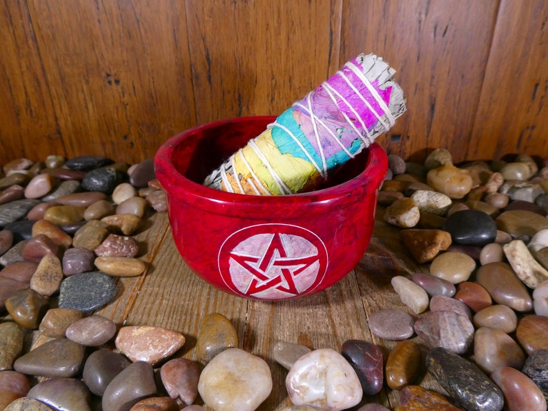 May include: A red, round, stone bowl with a white pentagram design. A white sage smudge stick with rainbow coloured string is inside the bowl. The bowl is sitting on a wooden surface with small, colourful stones around it.