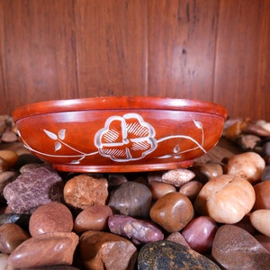 May include: A red wooden bowl with a white floral design sits on a bed of smooth, brown, gray, and white stones.