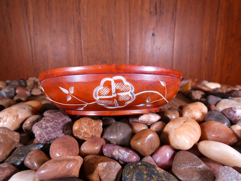 May include: A red wooden bowl with a white floral design sits on a bed of smooth, brown, gray, and white stones.