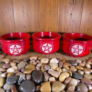 May include: Three red soapstone bowls with a white pentagram design. The bowls are sitting on a bed of small, smooth stones.