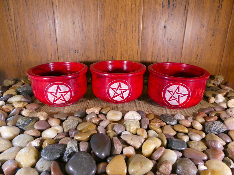 May include: Three red soapstone bowls with a white pentagram design. The bowls are sitting on a bed of small, smooth stones.