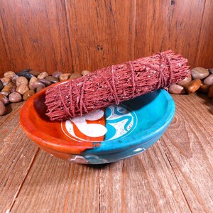 May include: A red sage smudge stick in a blue and orange ceramic bowl with an Om symbol. The bowl is surrounded by small brown and tan stones.