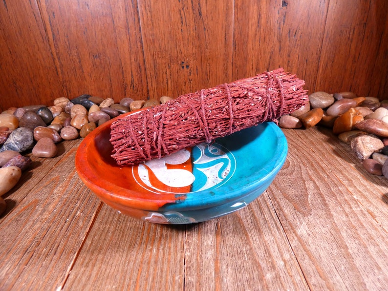 May include: A red sage smudge stick in a blue and orange ceramic bowl with an Om symbol. The bowl is surrounded by small brown and tan stones.