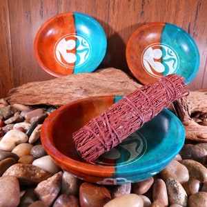 May include: Three teal and brown round bowls with a white Om symbol design. A bundle of red sage is resting in one of the bowls. The bowls are sitting on a bed of small brown and gray stones.