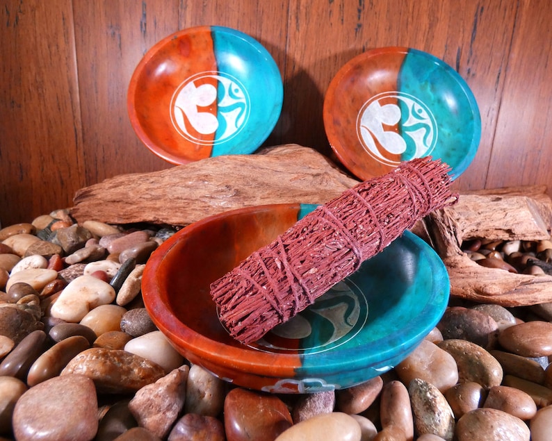 May include: Three teal and brown round bowls with a white Om symbol design. A bundle of red sage is resting in one of the bowls. The bowls are sitting on a bed of small brown and gray stones.