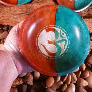 May include: A wooden bowl with a teal and red finish. The bowl has a white Om symbol carved into the surface.