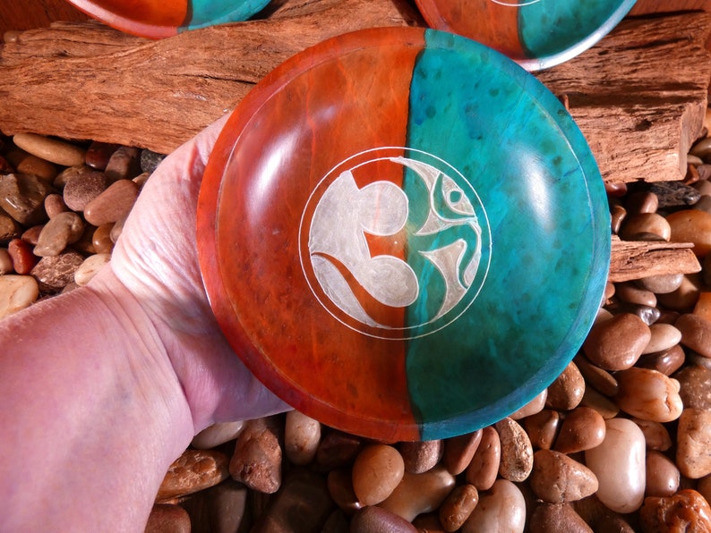 May include: A wooden bowl with a teal and red finish. The bowl has a white Om symbol carved into the surface.