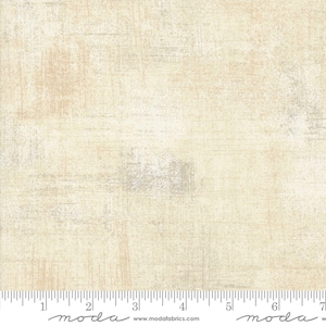 May include: A close-up of a beige fabric with a subtle textured pattern. The fabric has a slightly distressed look, with hints of grey and white throughout. The fabric is measured in inches with the text "moda" printed on the ruler.