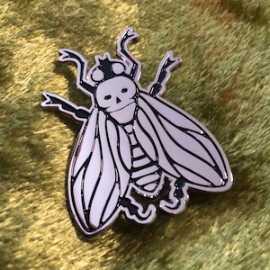 May include: A silver-colored enamel pin in the shape of a fly with a skull-shaped head. The fly has black details outlining the wings, body segments, and legs. The pin is set against a textured, olive-green background.