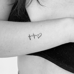 Puede incluir: Una foto en blanco y negro de un brazo con un tatuaje de una cruz y un latido del corazón con un corazón al final.