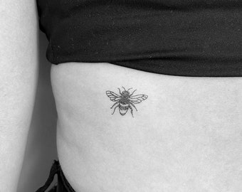 May include: A black and white tattoo of a bee on a person's lower abdomen.