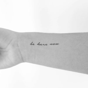 以下が含まれることがあります： 「be here now」と書いた、腕のタトゥーの白黒写真です。