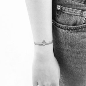 Puede incluir: Un delicado tatuaje de línea de dibujo de un símbolo egipcio antiguo en la muñeca de una persona.