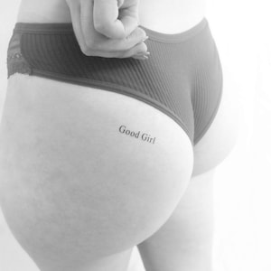 Puede incluir: Imagen en escala de grises de la parte baja de la espalda y las nalgas de una persona, con ropa interior rojo oscuro. La frase "Good Girl" está tatuada en la piel. Se ve una mano, que oscurece parcialmente la parte superior de la ropa interior.