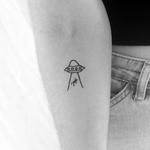 May include: A black and white tattoo of a UFO with a beam of light shining downwards. The UFO has a rounded top with several small circles. The beam of light is shining on a small figure.