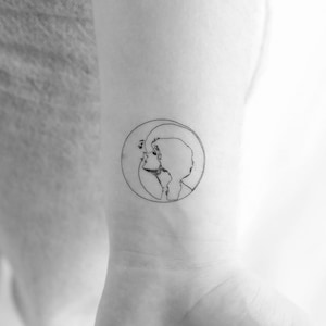Op de afbeelding: Een tattoo-ontwerp met een lijntekening van een vrouwengezicht met een halve maan achter haar hoofd.