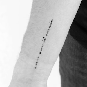 May include: A black and white photo of a person's arm with a tattoo that reads "amor vincit omnia" in cursive.