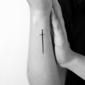 Può includere: Un piccolo tatuaggio a inchiostro nero raffigurante una spada sottile e dritta, posizionata verticalmente sull'avambraccio. Il disegno presenta una guardia semplice e una lama appuntita, realizzato con linee nere sottili e nette.