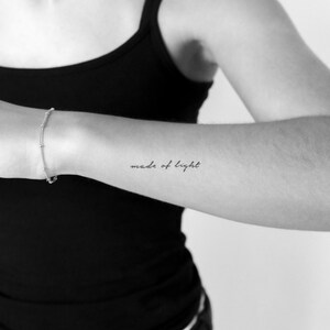 May include: A black and white image of an arm with a small silver bracelet and a tattoo that reads "made of light" in cursive script. The arm is against a black background.