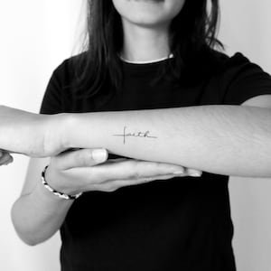 Op de afbeelding: Zwart-wit afbeelding van een pols met een kleine tatoeage. De tatoeage is het woord "faith" geschreven in een sierlijk, cursief schrift. De arm is omhoog gehouden en de persoon draagt een zwart shirt.