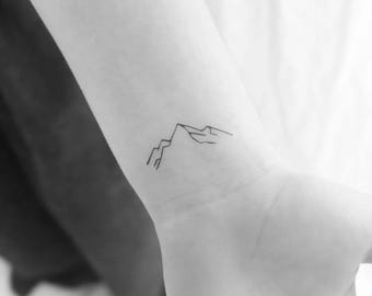 May include: A simple line drawing tattoo of a mountain range on a person's wrist.