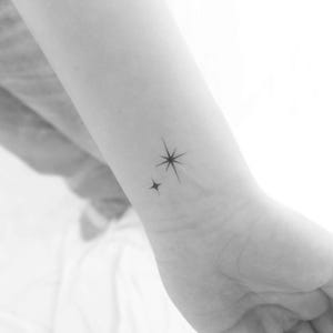 Peut inclure: Un petit tatouage simple de deux étoiles sur le poignet d'une personne. La plus grande étoile a cinq branches et la plus petite étoile a quatre branches.