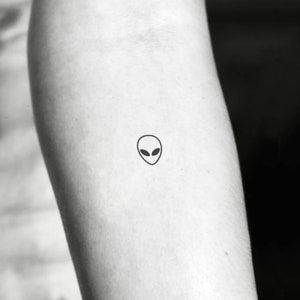 May include: A simple black line drawing of an alien head with large eyes on a person's arm.