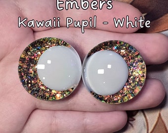 May include: Two white kawaii doll eyes with a gold glitter rim. The eyes have a white pupil and are labeled "Embers Kawaii Pupil - White".