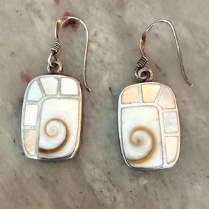 May include: A pair of rectangular drop earrings featuring a central spiral shell inlay surrounded by white mother-of-pearl segments set in a silver-toned metal frame. Each earring hangs from a curved silver-toned wire hook for pierced ears.