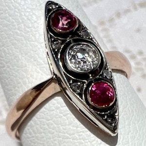 May include: A vintage ring with a gold band and a silver setting. The setting features a central diamond and two red gemstones. Small diamonds are set around the gemstones.