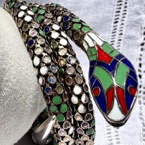 May include: A coiled snake bracelet featuring a silver-tone metal body with a mesh-like texture filled with small circular inlays in white, green, and blue. The head is decorated with vibrant red, blue, green, and white enamel geometric patterns.