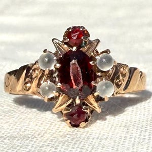 May include: A gold-toned ring featuring a large, oval-shaped, deep red gemstone at its center, surrounded by smaller red and white gemstones. The ring's band has an ornate, textured design, and the setting has a detailed, vintage style.