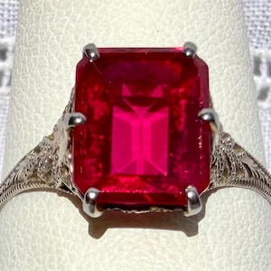 May include: A silver ring featuring a large, rectangular, vibrant red gemstone. The gemstone is the focal point, set in a detailed, filigree-style band. The ring's design is classic and elegant, with a vintage aesthetic.