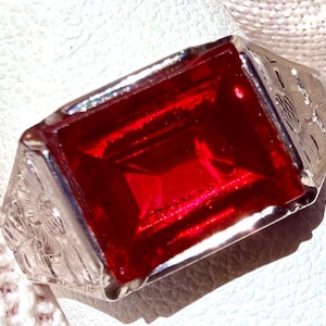 May include: A silver ring with a large, rectangular, deep red gemstone. The ring's band features intricate engraved floral patterns on the sides. The gemstone is set in a square bezel, creating a bold and eye-catching design.
