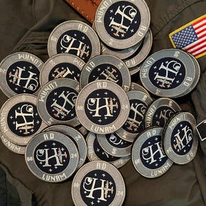 May include: A pile of circular embroidered patches. Each patch displays a stylized "H" with an airplane, encircled by text including "LUNAM", "AD", and numbers. The patches are mainly navy blue, grey, and white, with red accents. The patches are on a green background.