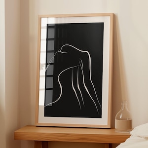 May include: A black and white line art print of a person's silhouette in a wooden frame. The print is hanging on a wall above a wooden table.