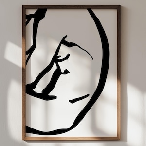 May include: Abstract black and white art print in a wooden frame. The artwork features bold, minimalist lines forming a face-like shape against a white background. The frame is a warm, natural wood tone, adding a touch of elegance.