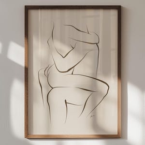 Puede incluir: Una impresión artística enmarcada con un dibujo lineal minimalista de dos figuras abrazándose. La obra de arte está realizada en una línea negra continua sobre un fondo crema. El marco es de madera.