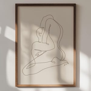 Op de afbeelding: Een minimalistische kunstprint met lijst. Het kunstwerk toont twee figuren in een omhelzing, weergegeven in een doorlopende zwarte lijn op een crèmekleurige achtergrond. De houten lijst voegt warmte toe aan het moderne ontwerp.