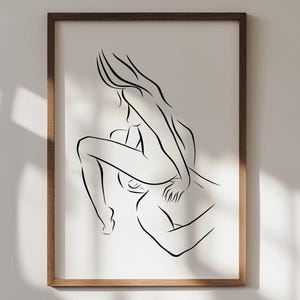 May include: A framed art print featuring a minimalist line drawing of two figures in an embrace. The artwork is rendered in black lines on a white background, encased in a brown wooden frame. The image captures a moment of intimacy.