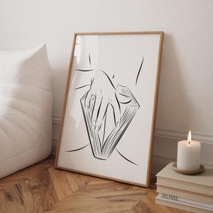 May include: A framed art print featuring a minimalist line drawing of a hand holding an open book. The artwork is in black and white, with a wooden frame. A lit candle and stacked books are in the foreground.