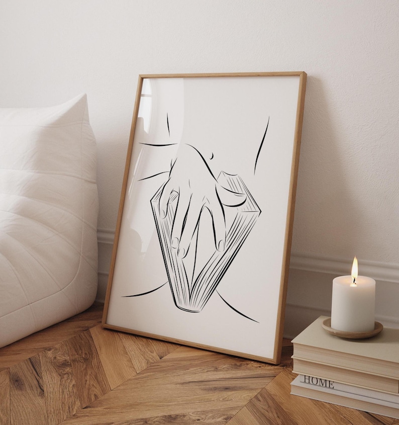 May include: A framed art print featuring a minimalist line drawing of a hand holding an open book. The artwork is in black and white, with a wooden frame. A lit candle and stacked books are in the foreground.