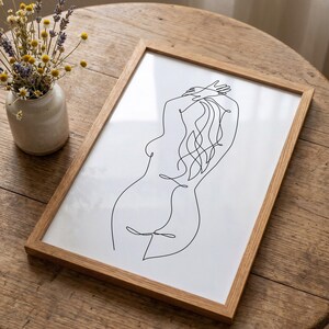 Puede incluir: Una impresi&oacute;n enmarcada con un dibujo lineal minimalista de la silueta de una persona. La obra de arte est&aacute; en negro sobre un fondo blanco, dentro de un marco de madera natural. La imagen est&aacute; sobre una mesa de madera, con un peque&ntilde;o jarr&oacute;n de flores secas.