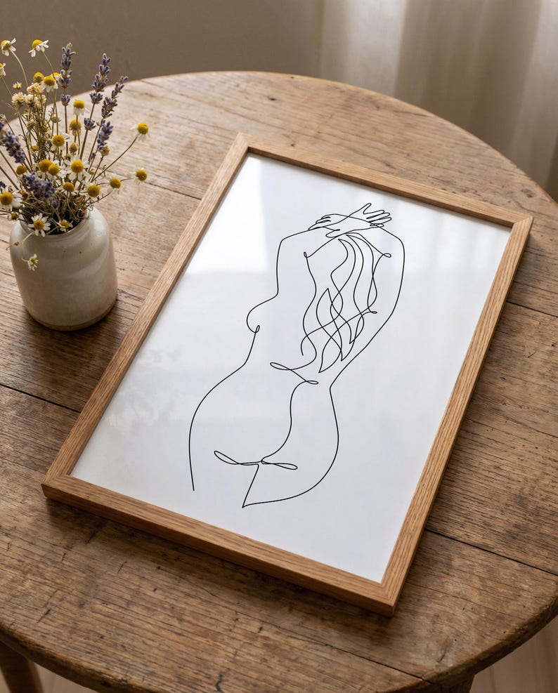 Puede incluir: Una impresi&oacute;n enmarcada con un dibujo lineal minimalista de la silueta de una persona. La obra de arte est&aacute; en negro sobre un fondo blanco, dentro de un marco de madera natural. La imagen est&aacute; sobre una mesa de madera, con un peque&ntilde;o jarr&oacute;n de flores secas.
