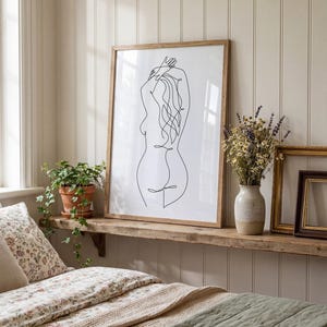 Puede incluir: Una impresi&oacute;n art&iacute;stica enmarcada con un dibujo lineal minimalista de una figura femenina. La obra de arte est&aacute; en un marco de madera natural y descansa sobre una estanter&iacute;a de madera r&uacute;stica. Tambi&eacute;n hay una maceta y un jarr&oacute;n con flores secas.