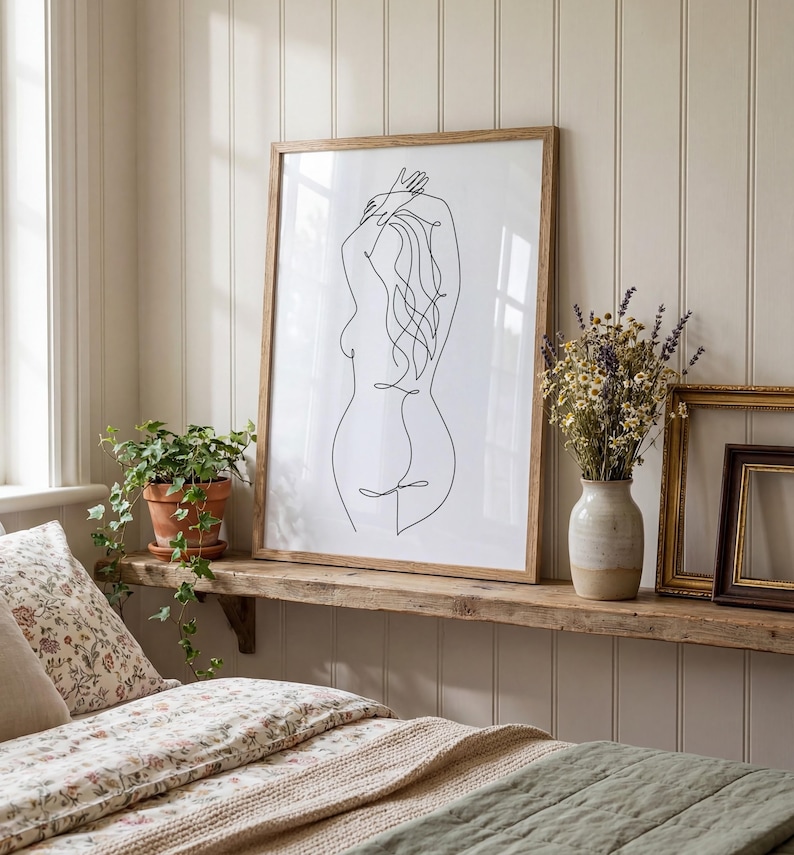 Puede incluir: Una impresi&oacute;n art&iacute;stica enmarcada con un dibujo lineal minimalista de una figura femenina. La obra de arte est&aacute; en un marco de madera natural y descansa sobre una estanter&iacute;a de madera r&uacute;stica. Tambi&eacute;n hay una maceta y un jarr&oacute;n con flores secas.